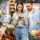Smiling Millennial Couple Shopping Groceries In The Hypermarket, Two Happy Consumers Buying Food Walking With Cart In Wholesale Shop, Choosing Eco Vegetables And Fruits, Checking Label And Expiry Date